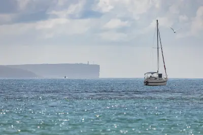 Sailing boat on the waters near Currambene Creek with the sheer cliffs and lighthouse of Point Perpendicular visible in the distance.