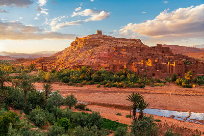 Sunset over Aït Benhaddou in Morocco showing the ancient earthen ksar, palm oasis, and surrounding desert landscape.