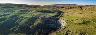 Aerial panoramic view of Malham Cove's curved limestone amphitheatre, surrounded by green hills and dry-stone walls in the Yorkshire Dales.