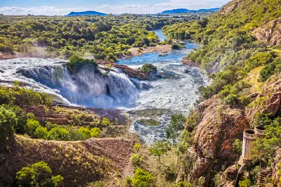 View of water cascading from Hartbeespoort Dam into the Crocodile River below, surrounded by rocky cliffs, dense vegetation and the Magaliesberg foothills.