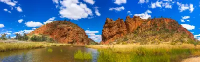 A wide panoramic landscape shows the Glen Helen Gorge waterhole flowing through a grassy desert oasis, flanked by massive, rugged orange cliffs under a bright blue sky with scattered clouds.