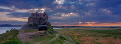 Panoramic sunset view of Lindisfarne Castle on its rocky outcrop, with dramatic purple-blue clouds, golden light, coastal paths and tidal flats on Holy Island, Northumberland.