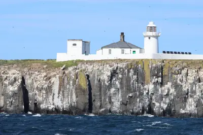White lighthouse and adjoining buildings on Inner Farne Island, perched above steep rocky cliffs with seabirds and the North Sea below, Northumberland.