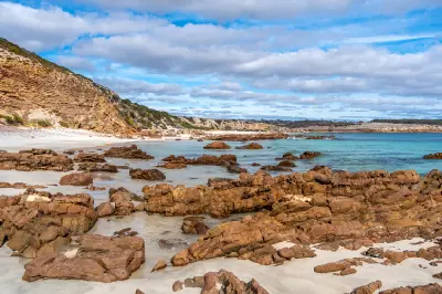 Reddish-brown coastal rocks scattered across white sand beside turquoise water and layered cliffs at Stokes Bay, Kangaroo Island, South Australia.