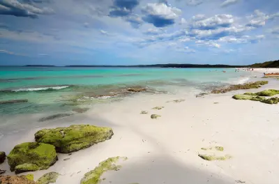 White sand, moss-covered shoreline rocks and clear turquoise water at Hyams Beach beneath a cloud-filled sky.