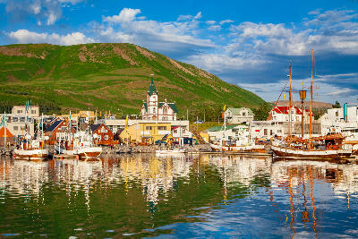 Húsavík harbour in North Iceland at sunset, showing boats reflected in calm water with the town and surrounding green hills in the background.