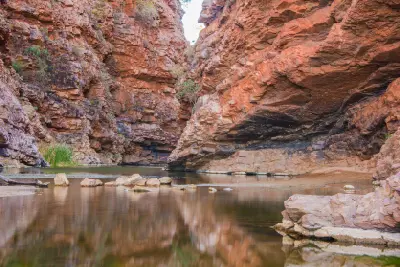 Striated orange-red rock formations reflecting perfectly across the still surface of a scenic outback pool at Simpsons Gap, or Rungutjirpa.