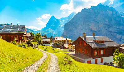 Mürren village with chalets and a winding path across green slopes, overlooking steep cliffs and high alpine peaks in the Swiss Alps.