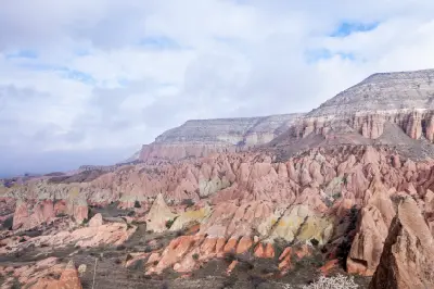 Panoramic view of Red Valley in Cappadocia, Turkey, with colourful red, pink and cream volcanic ridges beneath layered cliffs and a cloud-filled sky.