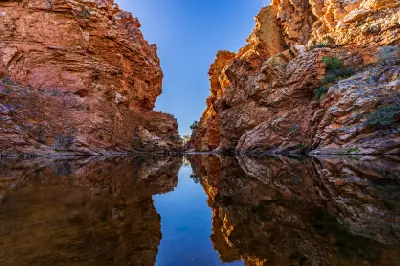 Sheer red-orange rock walls of a narrow outback gorge create a perfect reflection on the calm, glassy surface of the nature waterhole landscape.
