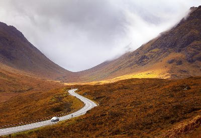 Winding road through the Scottish Highlands in autumn, with golden foliage and rolling hills under changing light.