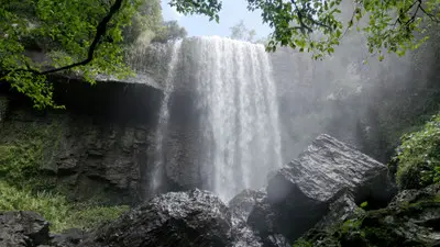 Low-angle view of Zillie Falls cascading over a dark rock cliff, with large foreground boulders, mist and tropical vegetation in Queensland, Australia.