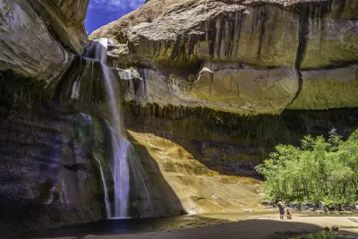 Lower Calf Creek Falls plunges from a towering sandstone alcove into a pool, with visitors resting on the sandy shore below.