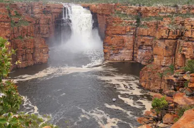 King George Falls plunges between towering red sandstone cliffs into the broad gorge below, with small boats on the river providing scale.