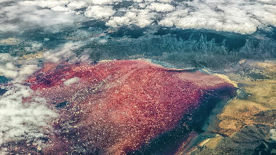 Aerial view of Lake Natron showing bright red alkaline waters, salt flats, and the Great Rift Valley escarpment beneath scattered clouds in northern Tanzania.