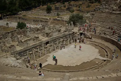 Elevated view inside the Great Theatre of Ephesus showing tourists among the ancient stage ruins, stone columns, and curved tiers of seating.