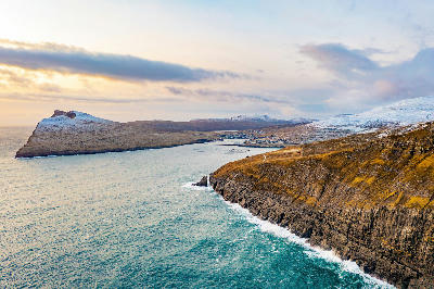 Aerial view of Sandavágur village on Vágar Island, showing rocky sea cliffs, a sheltered bay, fish farms offshore, and snow-covered hills in the Faroe Islands.