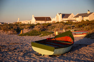Colourful fishing boat on a sandy beach in Paternoster at sunset, with whitewashed village houses and coastal vegetation in the background.