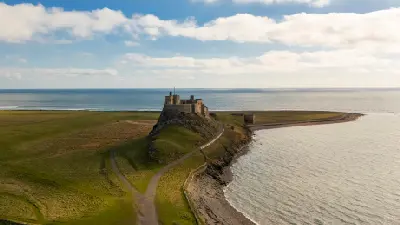 Aerial view of Lindisfarne Castle on Holy Island, Northumberland, showing the stone fortress on a rocky hill, winding approach path, grassy coastline and surrounding sea.