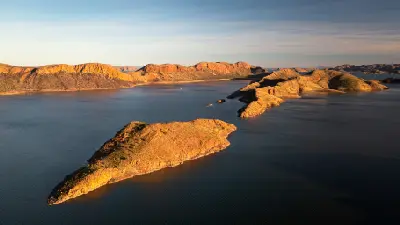 Golden evening light illuminates rugged rocky islands surrounded by the deep blue waters of Lake Argyle in the Kimberley.