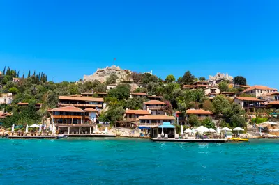 Kaleköy in Kekova, Antalya, Turkey, with waterfront houses, lush hillside vegetation, turquoise sea, and historic Simena Castle overlooking the village.