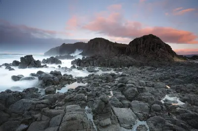 Rugged volcanic rock formations and tide pools at Minnamurra Point, with waves breaking beneath pink clouds at dusk.