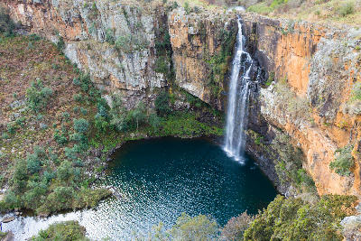 Berlin Falls cascading into a dark circular plunge pool surrounded by orange rock cliffs and indigenous vegetation in Mpumalanga.