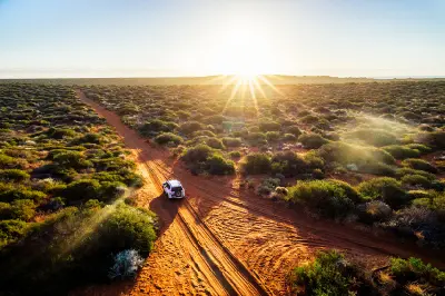 Aerial view of a four-wheel drive travelling along red sand tracks through coastal scrub in golden evening light.