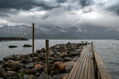 Wooden pier in Mefjordvær fjord with rocky shoreline and cloud-covered mountains on Senja island, Norway