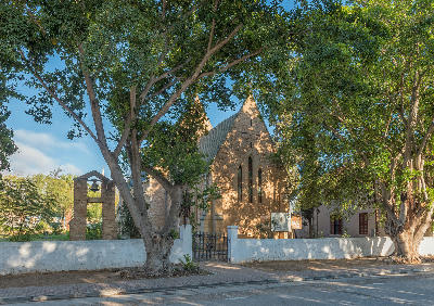 Historic Anglican Church in Clanwilliam, Western Cape, with a stone belfry and bell surrounded by large shade trees along a quiet town street.