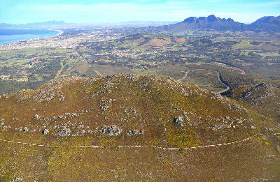 High aerial landscape of Sir Lowry’s Pass with rocky fynbos ridges overlooking Somerset West, Strand, False Bay, and distant Table Mountain in the Western Cape, South Africa.