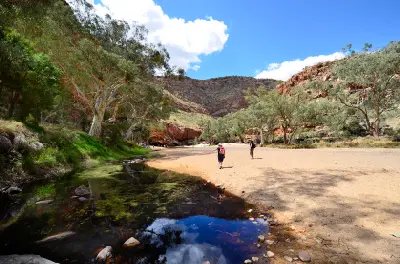 Visitors cross the sandy floor of Ormiston Gorge beside a shallow waterhole, river gums and rugged red cliffs.