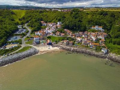 Aerial photograph of Runswick Bay showing hillside cottages, a sandy beach, stone seawall and wooded coastal slopes in North Yorkshire, England.