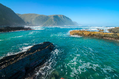Turquoise ocean and rocky shoreline beneath coastal mountains along the Tsitsikamma Coast in South Africa.