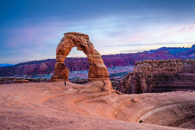 Delicate Arch with person standing beneath it at sunset with La Sal Mountains in the background in Arches Utah