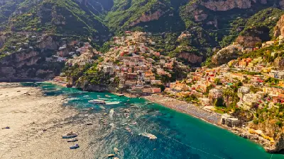 Aerial view of the Amalfi Coast, with colourful hillside houses above a turquoise sea, boats along the shoreline, and steep green cliffs in southern Italy.