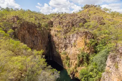 High-angle view of Tolmer Falls dropping into a dark plunge pool beneath towering sandstone cliffs, with green woodland and rocky hills in Litchfield National Park, Northern Territory, Australia.
