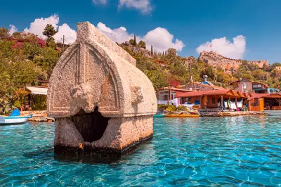 Ancient Lycian sarcophagus standing in clear turquoise water at Kaleköy, Turkey, with waterfront houses and Simena Castle on the hillside behind.