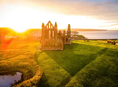 Elevated sunset view of Whitby Abbey's Gothic ruins and green grounds, overlooking Whitby town and the North Sea in North Yorkshire, England.