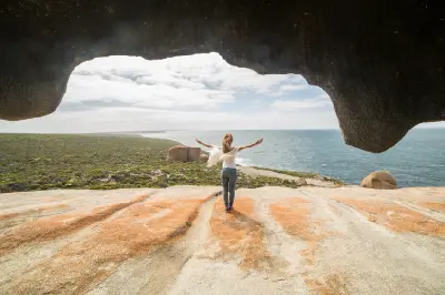 Woman standing with outstretched arms beneath a huge overhanging granite rock at Remarkable Rocks, overlooking the Southern Ocean on Kangaroo Island, South Australia.
