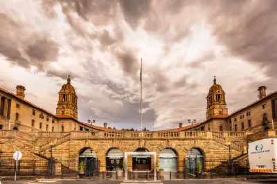 The Union Buildings in Pretoria beneath dramatic storm clouds, with the twin sandstone towers and central flagpole framed by a moody sky.