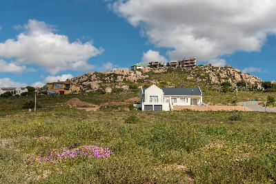 Wildflower-covered coastal vegetation and modern houses beneath granite hills in St Helena Bay, South Africa, under dramatic clouds and a blue sky.