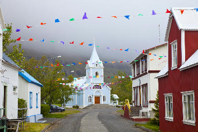 View down a narrow street toward the blue church in Seyðisfjörður, Eastfjords, Iceland, with traditional wooden houses and colorful flags overhead.
