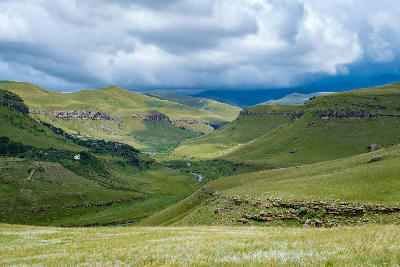 View across the Giant’s Castle region of the Central Drakensberg, showing green valleys, rolling mountains, rocky escarpments and a winding river beneath dramatic storm clouds in KwaZulu-Natal, South Africa.