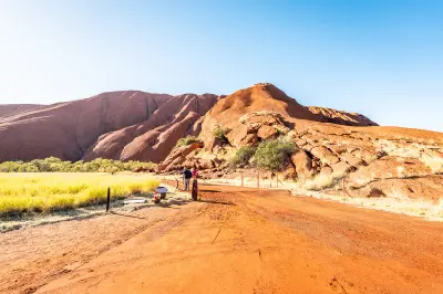 Visitors walking on a red-dirt trail beside the sandstone base of Uluru in Uluru-Kata Tjuta National Park, Australia.