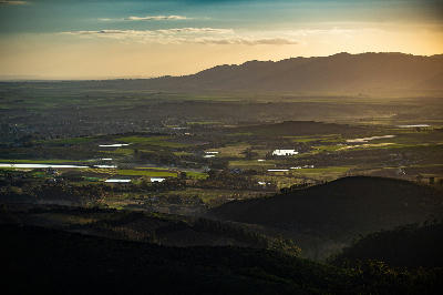 Golden sunset over the Cape Winelands near Wellington, South Africa, showing layered agricultural fields, farm dams, rolling valley terrain, and distant mountain silhouettes.