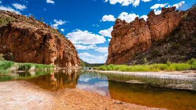 The towering orange rock cliffs of Glen Helen Gorge reflect perfectly in the calm, glassy waterhole below under a blue sky streaked with light clouds.