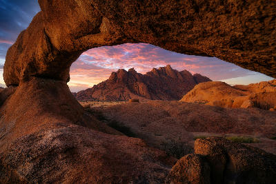 Natural granite rock arch framing the rugged peaks of Spitzkoppe at sunset, with glowing pink clouds and weathered granite formations across the desert landscape of central Namibia.