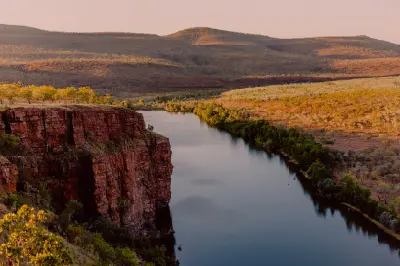 Warm evening light falls across a broad river, towering red cliffs and rugged ranges viewed from Branco’s Lookout at El Questro.