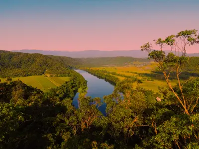 A calm river reflects the morning light as it curves through a dense tropical valley beneath a soft pink horizon.